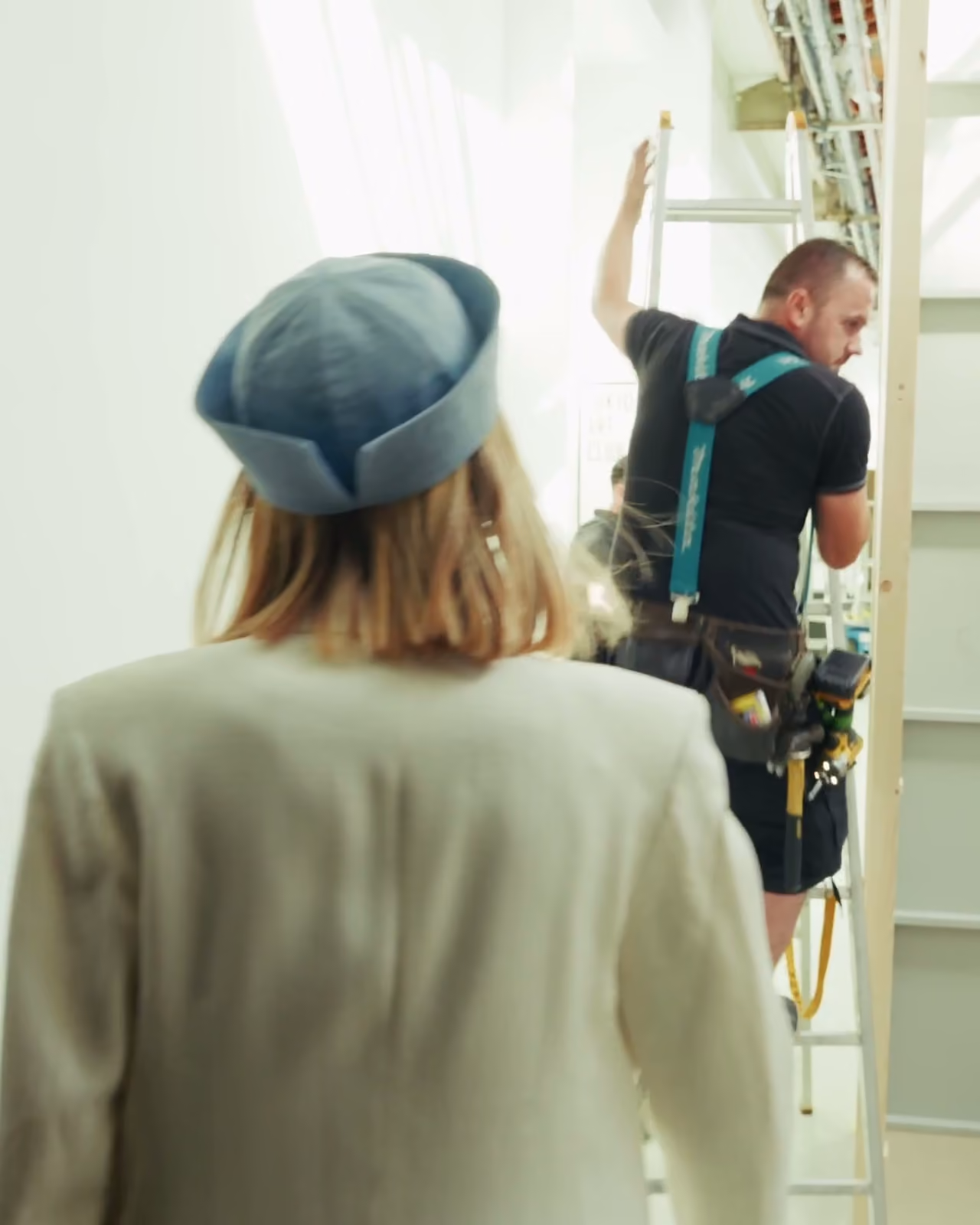 De dos, une jeune femme au chapeau bleu passe devant un technicien perché sur une échelle, en coulisses du défilé Istituto Marangoni