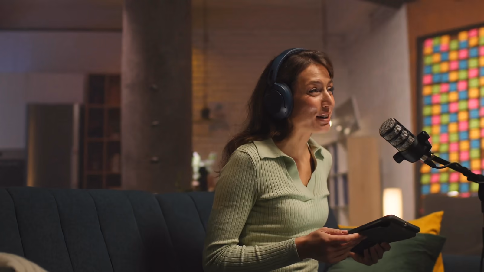 Une femme enregistre un podcast au micro, casque sur la tête, une tablette à la main.