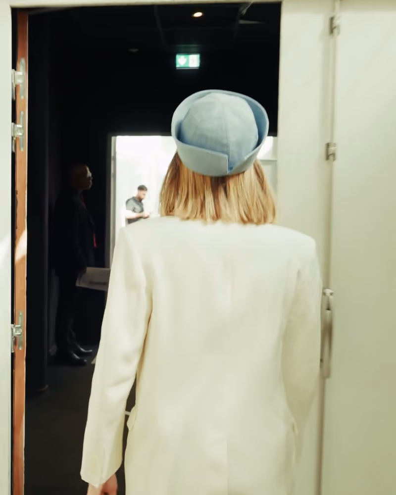 De dos, la jeune femme au chapeau bleu franchit une porte vers les coulisses du défilé